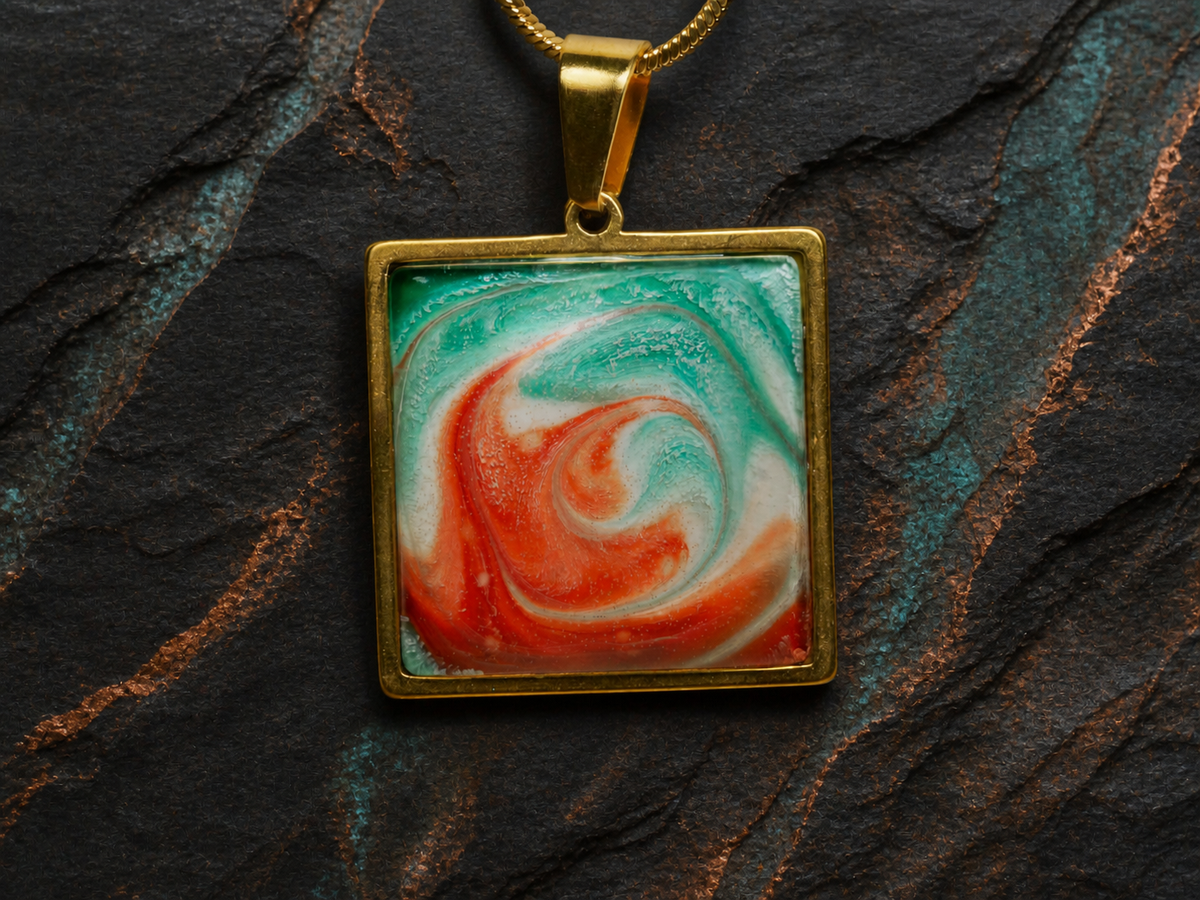 Close-up of square gold-plated pendant with turquoise and coral ink and resin artwork on slate