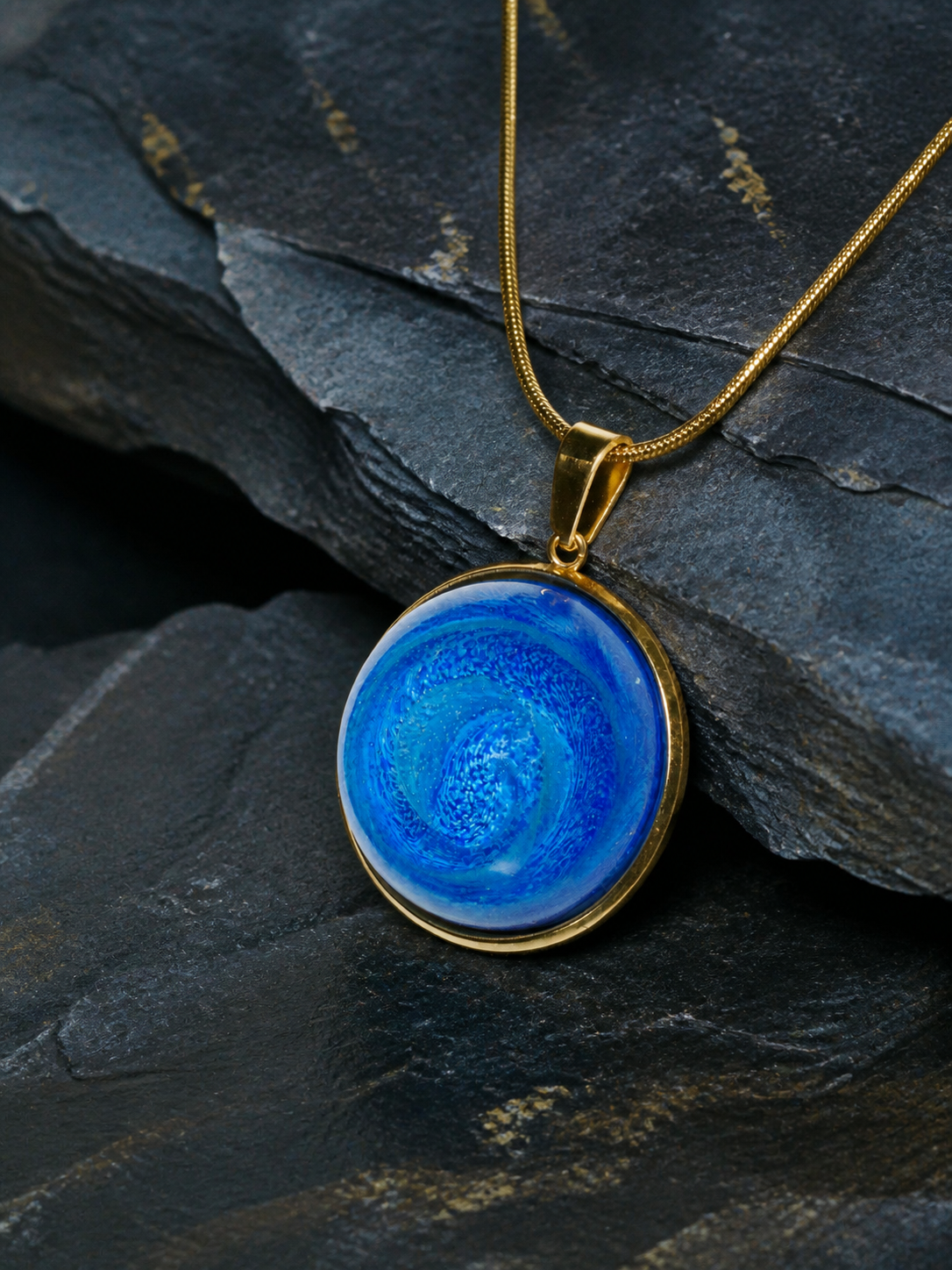 Close front view of the Tethys Gyre pendant, gold-plated bezel and snake chain on blue-grey shale