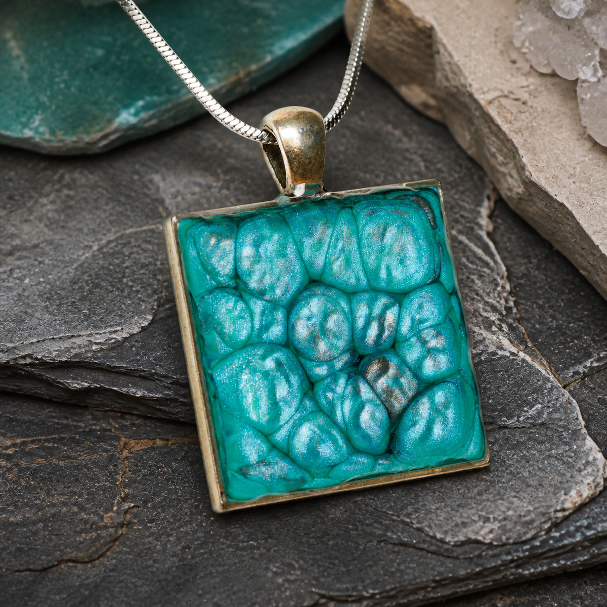 Close-up of silver square pendant with teal enamel-painted, resin-coated surface
