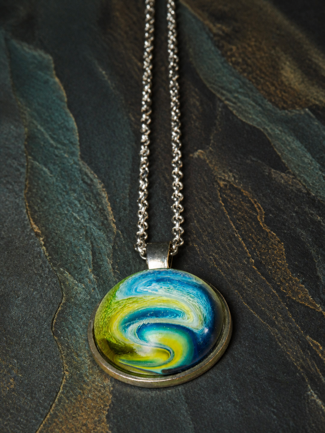 Verdant Gyre round pendant with silver chain arranged vertically on dark mineral stone